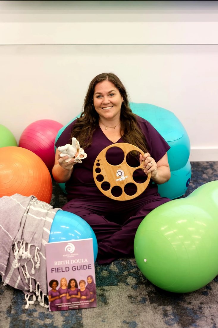 Madeline Harris LeBlanc sitting among birth balls, holding a model pelvis and a wooden dilation wheel, with the Birth Doula Field Guide beside her